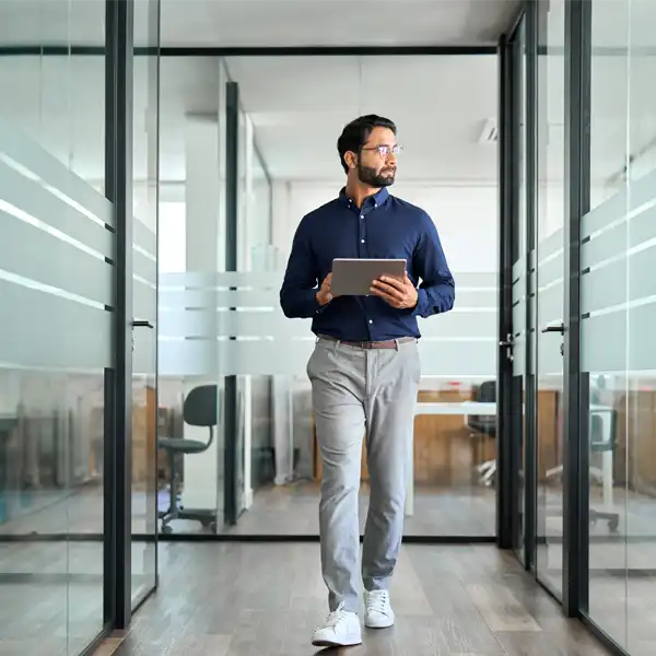 A man dressed in business casual walking and holding a digital device at the office