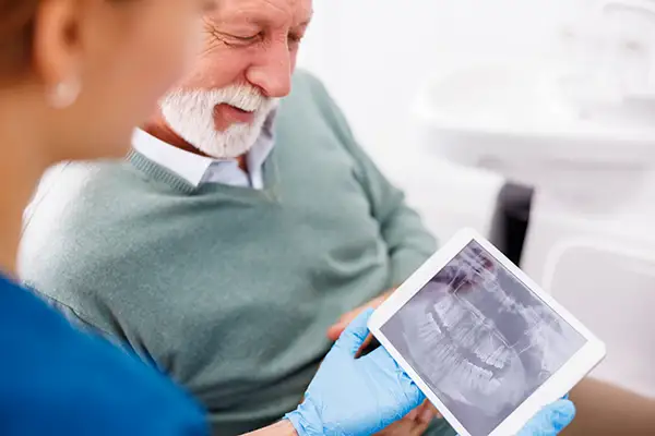 A dental assistant showing a digital x-ray to a senior patient on a tablet during a dental consultation.